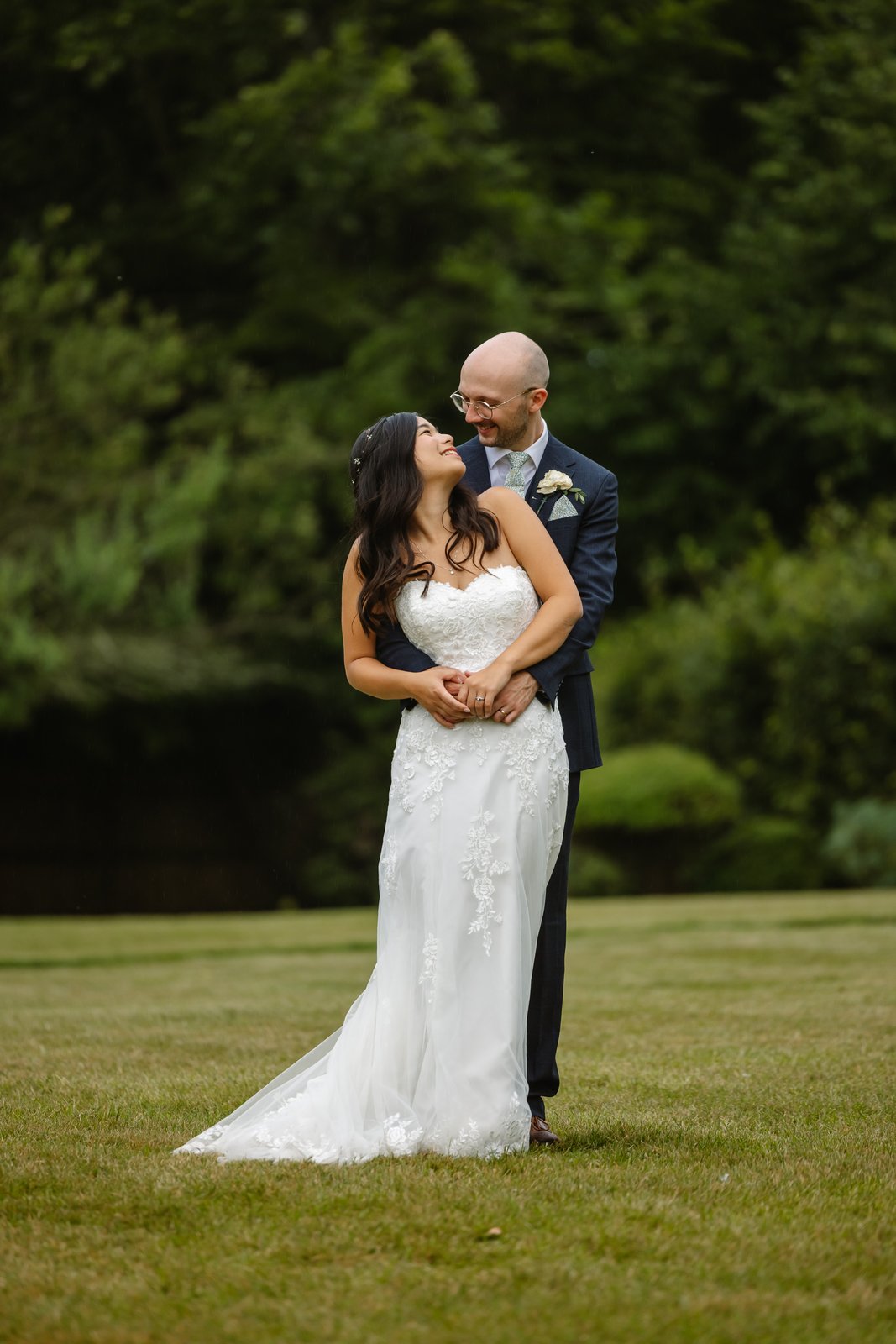 Bride and groom embracing on the open lawn at Worton Hall with trees behind.
