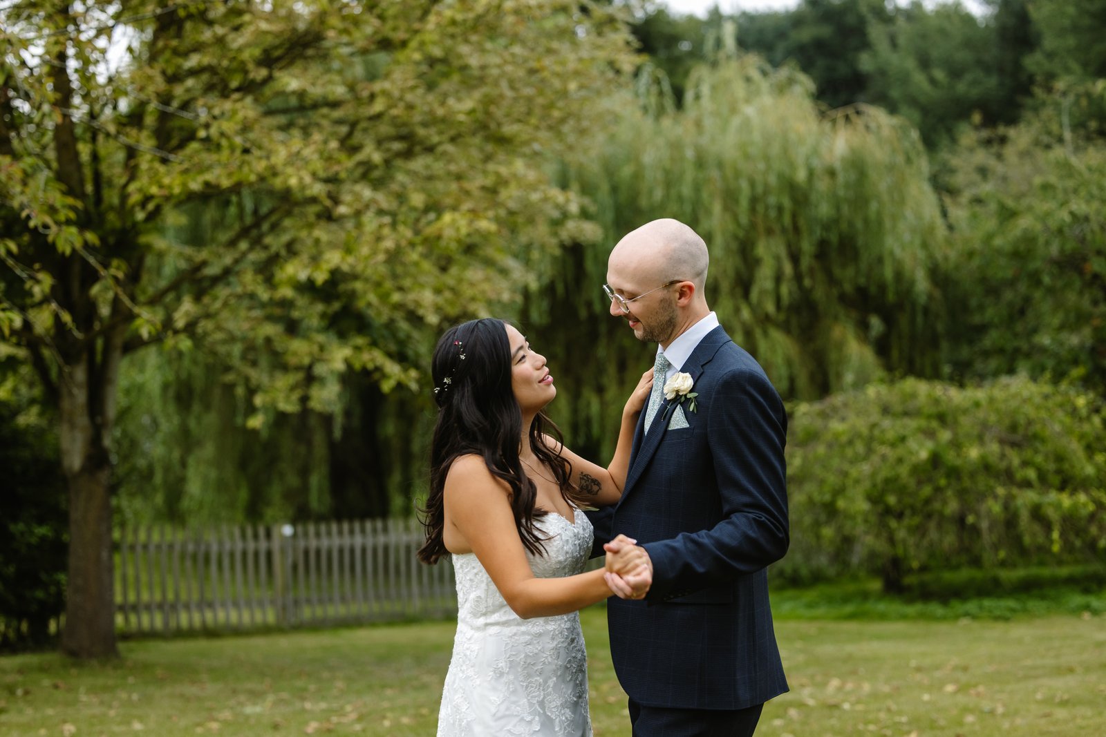 Bride and groom holding hands and dancing together beneath a willow tree.