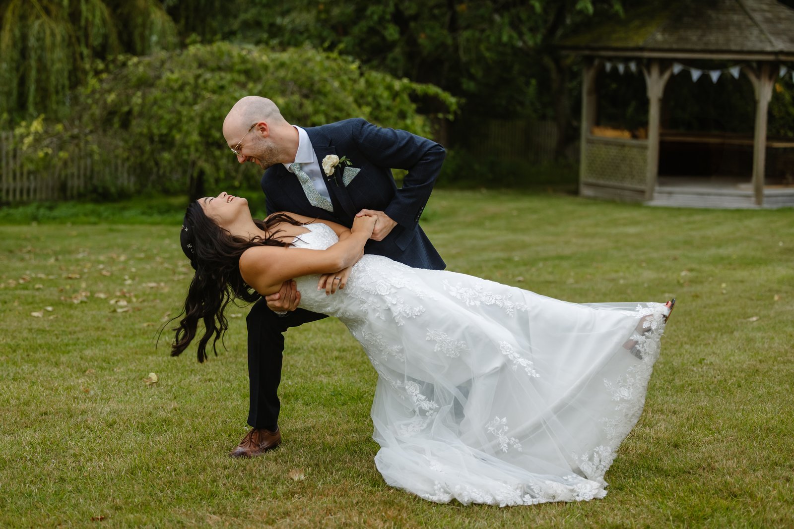 Groom dipping the bride backwards on the grass as she laughs, the gazebo behind them.
