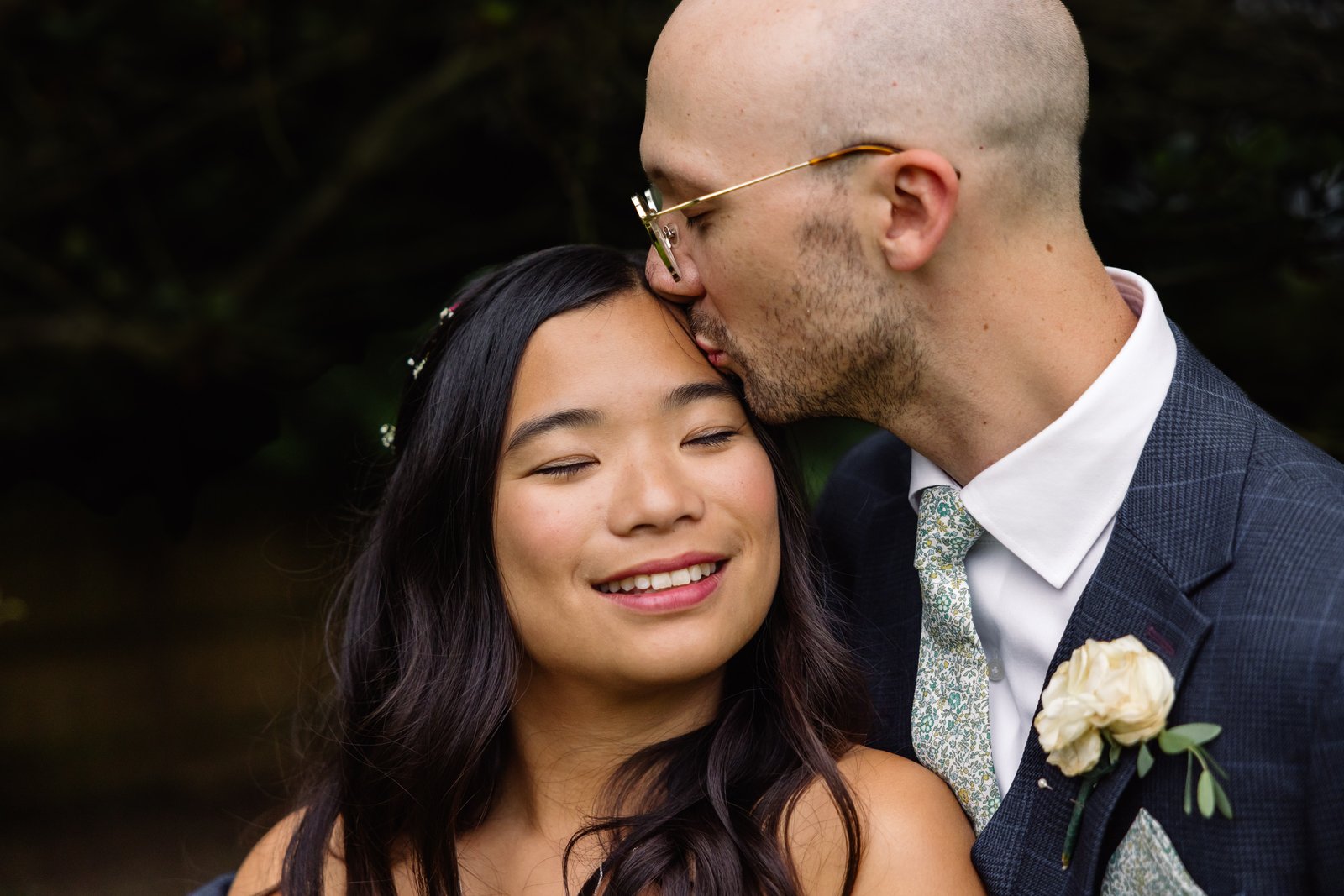 Groom kissing the bride's temple with the pond and trees behind them.