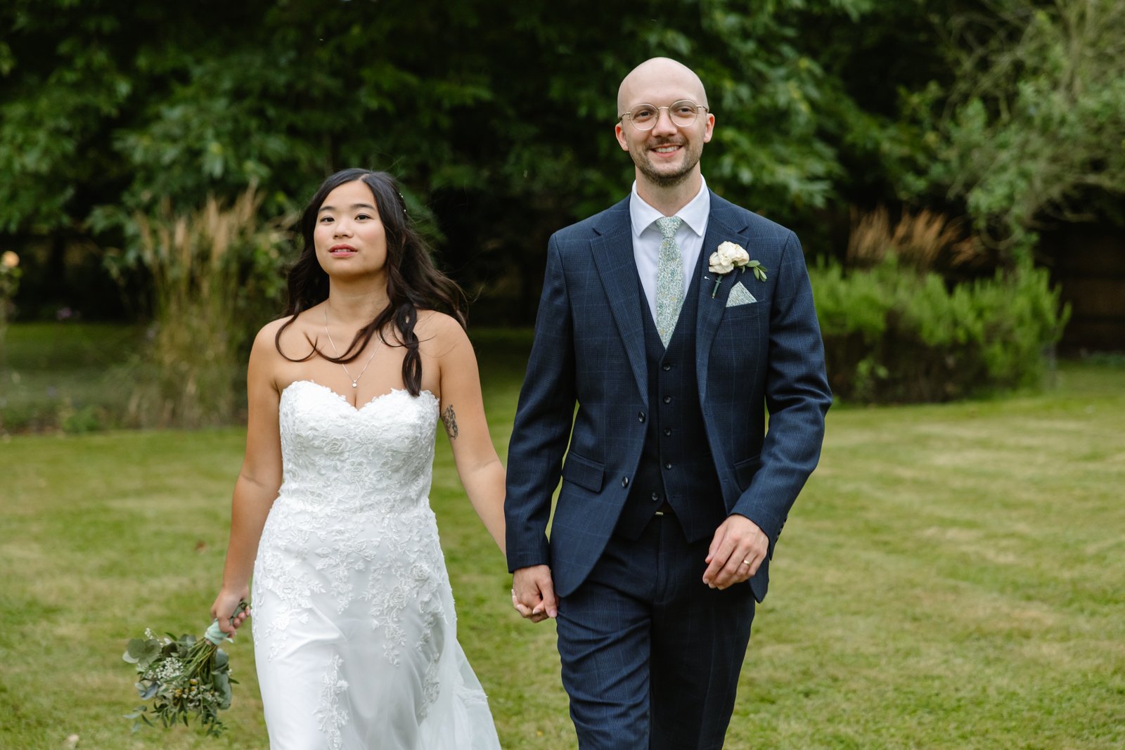 Bride and groom walking across the lawn at Worton Hall past tall ornamental grasses.
