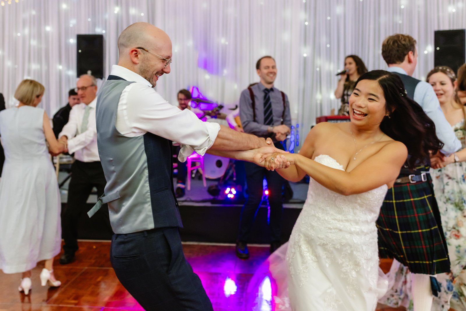 Bride dancing with the groom, his jacket off, on a crowded dance floor at the evening party.