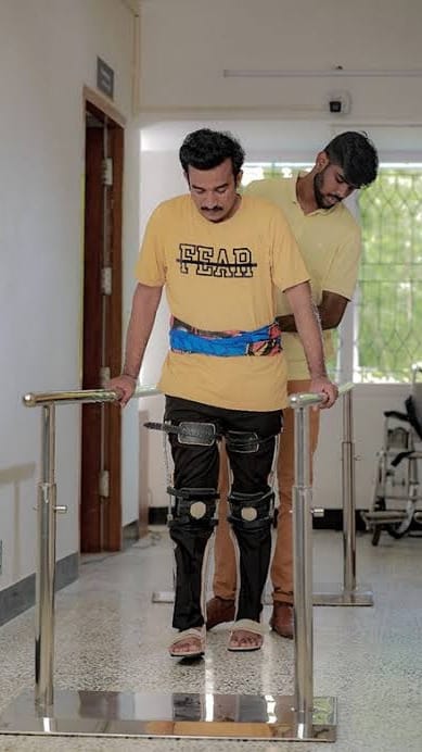 Patient wearing knee braces walking between parallel bars with a physiotherapist supporting him
