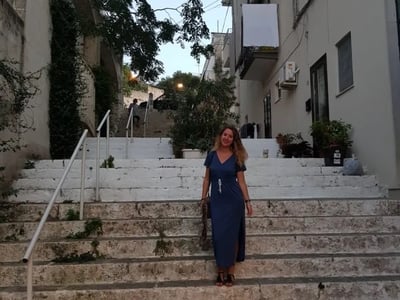 Woman in a blue dress descending stone stairs in Ostuni, surrounded by Mediterranean vegetation and historic buildings.