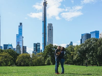 A couple kissing in Central Park, with modern skyscrapers in the background under a clear blue sky.