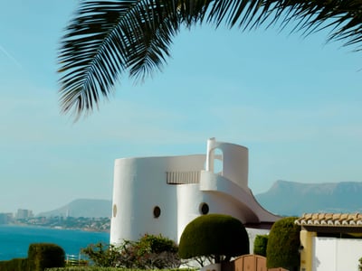Vue panoramique sur une maison moderne à Moraira, avec la mer Méditerranée en arrière-plan et des palmiers.