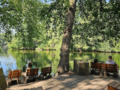 Two people sitting on wooden benches, facing a peaceful lake, surrounded by green trees at the Café am Neuen See.