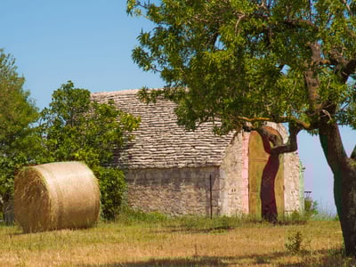 Grange en pierre typique de la région de Castellana Grotte, entourée d'arbres et de balles de foin dans un champ verdoyant.