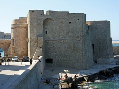 Ruines du Château de Charles V surplombant la plage, avec des visiteurs profitant du soleil et de la mer à proximité.
