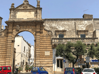 Majestic arch at the entrance of Piazza Manfredi, with historic buildings and Mediterranean vegetation.