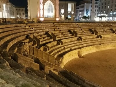 Amphithéâtre romain de Lecce illuminé la nuit, mettant en avant ses gradins historiques et l'architecture environnante.