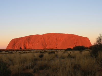 Uluru illuminated by the setting sun, with fields of vegetation around, an iconic symbol of Australia.