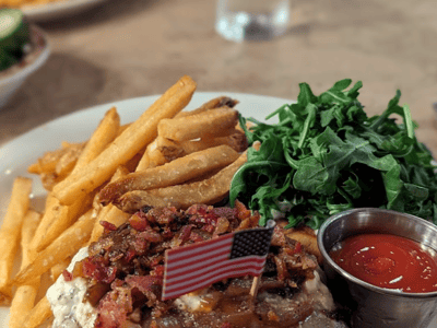 A burger topped with bacon and a small American flag, accompanied by golden fries and arugula, served at Ted's Montana Grill.