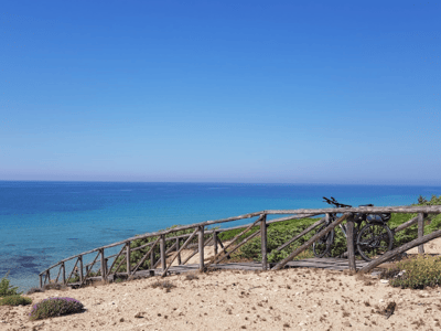 Vue côtier de Campomarino di Maruggio avec un sentier en bois menant à la mer turquoise sous un ciel ensoleillé.