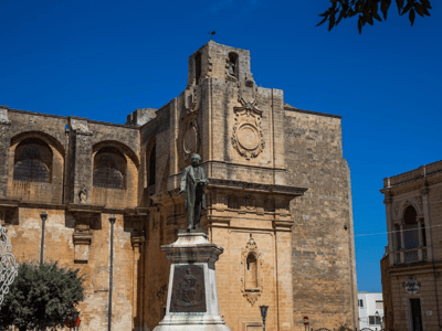 Statue of a man facing the Palazzo Gallone, a historic building with warm colors and a bright blue sky.
