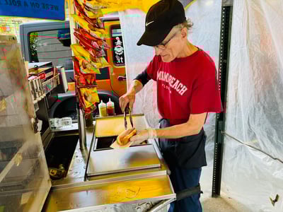 A vendor prepares a hot dog on Billy's cart, surrounded by bags of chips, in a lively street atmosphere.