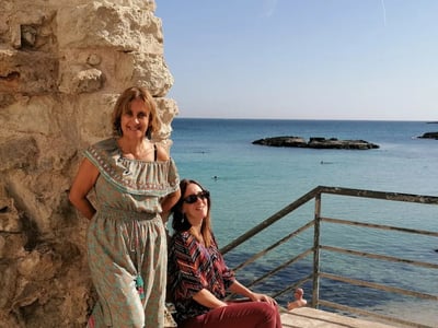 Deux femmes souriantes posent près des côtes pittoresques de Monopoli, avec une mer turquoise et des rochers en arrière-plan.