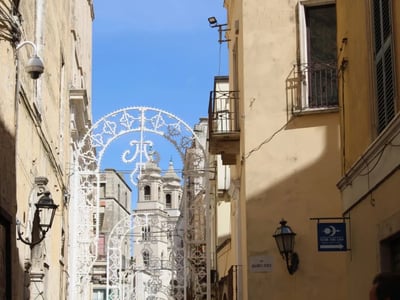 Vue d'une rue d'Altamura, avec un portail décoratif et la silhouette d'une église sous un ciel bleu.