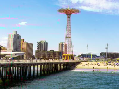 The famous Parachute Jump stands on the Coney Island pier, with modern buildings and the lively beach in the background.