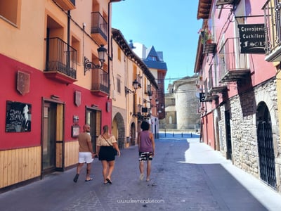 In the sunny streets of Jaca, three people stroll between colorful buildings, overlooking an ancient fortification.
