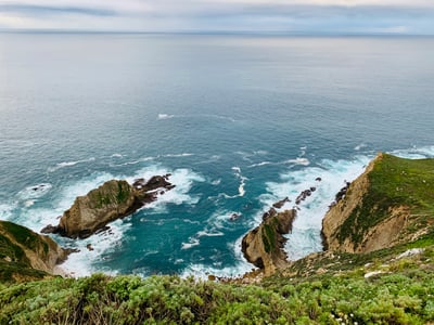 Vue panoramique de la côte de Big Sur, où les vagues frappent les rochers, entourée de verdure luxuriante et d'un ciel nuageux.