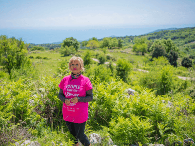 Femme en t-shirt rose, posant avec un paysage verdoyant et côtier en arrière-plan, lors du Festival des orchidées.