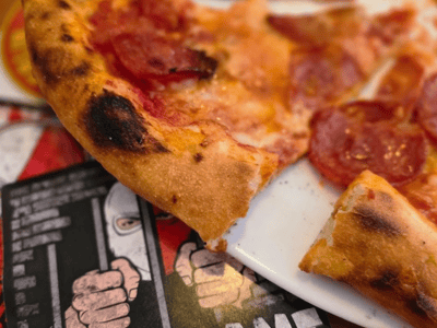 Détail d'une part de pizza au pepperoni au Salami Social Club, avec une carte amusante en arrière-plan.