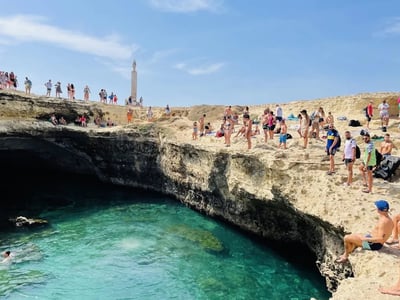 Gathering of bathers at the edge of the Caves of Poetry, with a turquoise sea below and a lighthouse in the background.