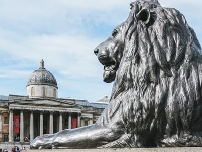 Bronze lion in Trafalgar Square, with the National Gallery in the background under a blue sky.