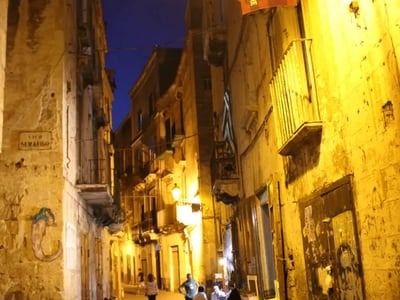 Illuminated alley of Taranto, showing ancient walls and residents chatting in a warm nighttime atmosphere.