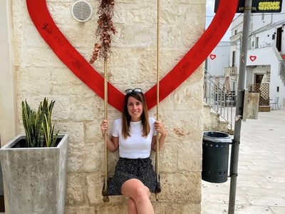 Woman sitting on a swing hanging from a large red heart in Cisternino, adding a romantic touch to the square.