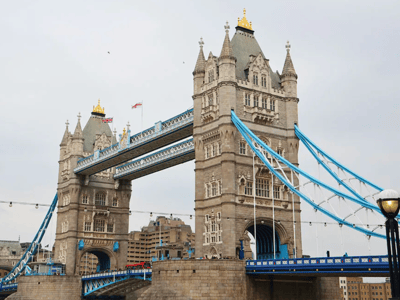 Tower Bridge in London, iconic with its majestic towers and blue cables under a cloudy sky.
