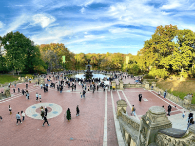 Crowd gathered around the Bethesda Fountain in Central Park, under a blue sky with colorful autumn foliage.
