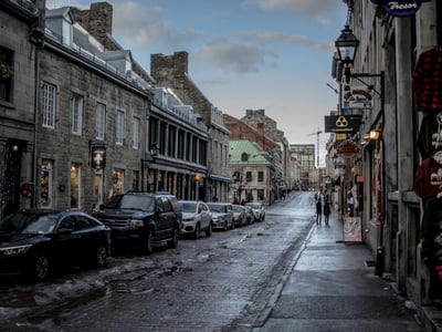 Paved street of Old Montreal, lined with historic buildings and cars, under a cloudy sky.