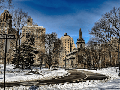 A winter landscape at Sheep Meadow, with majestic buildings and a blue sky, reflecting the beauty of Central Park.