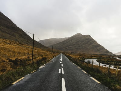 Winding and hilly road near Clifden, surrounded by brown hills and a peaceful river under a cloudy sky.