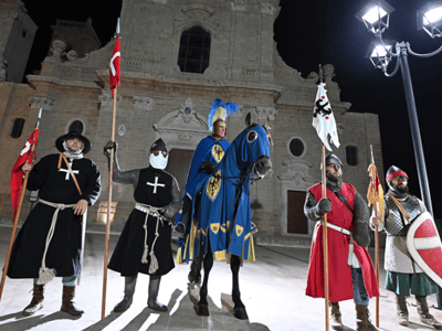 Five characters in medieval costumes standing in front of a church, representing the Palio of Oria in the evening.