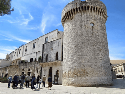 Touristes admirant la tour médiévale à Conversano, entourée de bâtiments historiques sous un ciel ensoleillé.