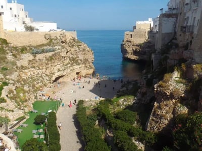 Lama Monachile beach nestled between cliffs, with bathers and white buildings in the background under a blue sky.