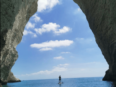 Surfer en paddle à travers l'arche naturelle des Grottes Marines, avec un ciel bleu et des vagues calmes en toile de fond.