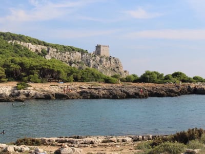 View of the Caves of Poetry, with a castle overlooking the sea and visitors walking on the rocky beach.
