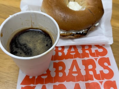 Un bagel garni avec de la crème et une tasse de café, servi sur un emballage coloré de Zabar’s, symbole de délices new-yorkais.