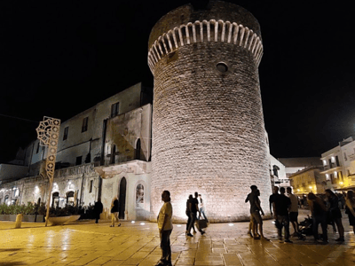 Tour médiéval de Conversano illuminé la nuit, montrant la tour en pierre et des visiteurs admirant son architecture historique.