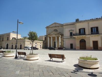 Calm place in Ugento with benches, trees, and a historic building in the background, under a sunny sky.