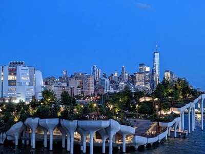 View of the illuminated Pier 57 Rooftop Park in the evening, with the iconic silhouette of Manhattan in the background.