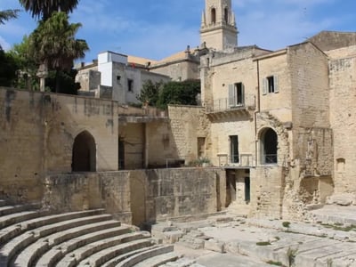 Amphithéâtre romain de Lecce, vestige historique entouré de bâtiments anciens et de palmiers, sous un ciel lumineux.
