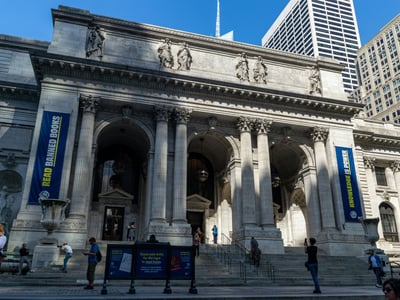 Majestic facade of the New York Public Library, with visitors ascending the steps under a blue sky.