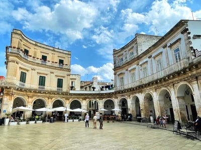 Plaza historique à Martina Franca avec des arches, des terrasses et un ciel bleu parsemé de nuages.