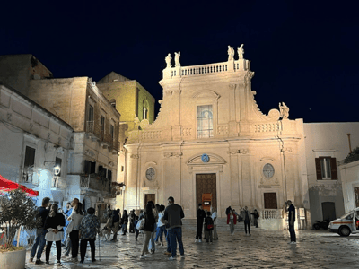 The illuminated façade of the Cathedral of Castellaneta, animated by evening visitors, reflects the local history.