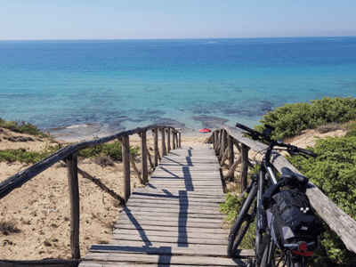 A wooden path leading to a peaceful beach at Campomarino di Maruggio, with a bike parked on the right.